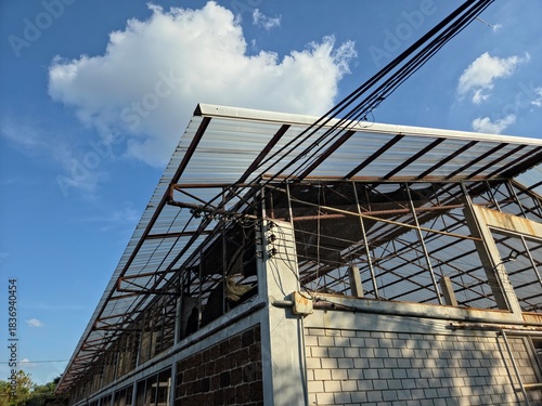 A low-angle shot of a large, long building with a metal frame and translucent roof partially covered by white brick and brown concrete walls against a blue sky with white clouds.