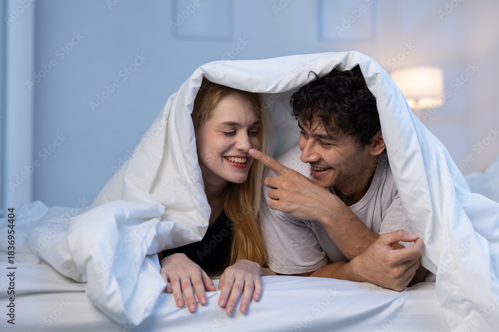 Fototapeta premium Happy couple playing under blanket in bed, expressing love, warmth, and joyful relationship moments. Perfect for romance, intimacy and lifestyle themes in cozy home.