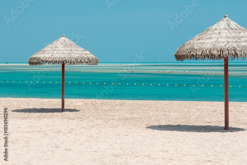 Sunny beach with two straw umbrellas over quiet turquoise water and soft sand, blue horizon