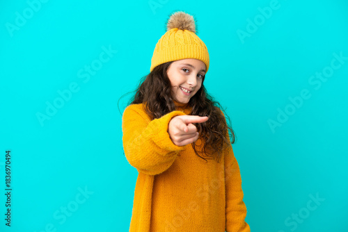 Little caucasian girl wearing winter hat isolated on blue background surprised and pointing front