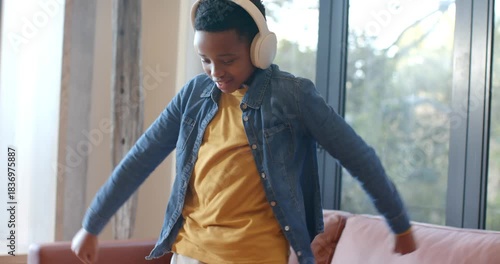 African American boy cueing music with white headphones, dancing in yellow T-shirt near glass door