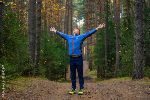 Young man standing on a forest trail with arms outstretched, breathing fresh autumn air and enjoying a peaceful, energizing moment of freedom and mindfulness while jogging
