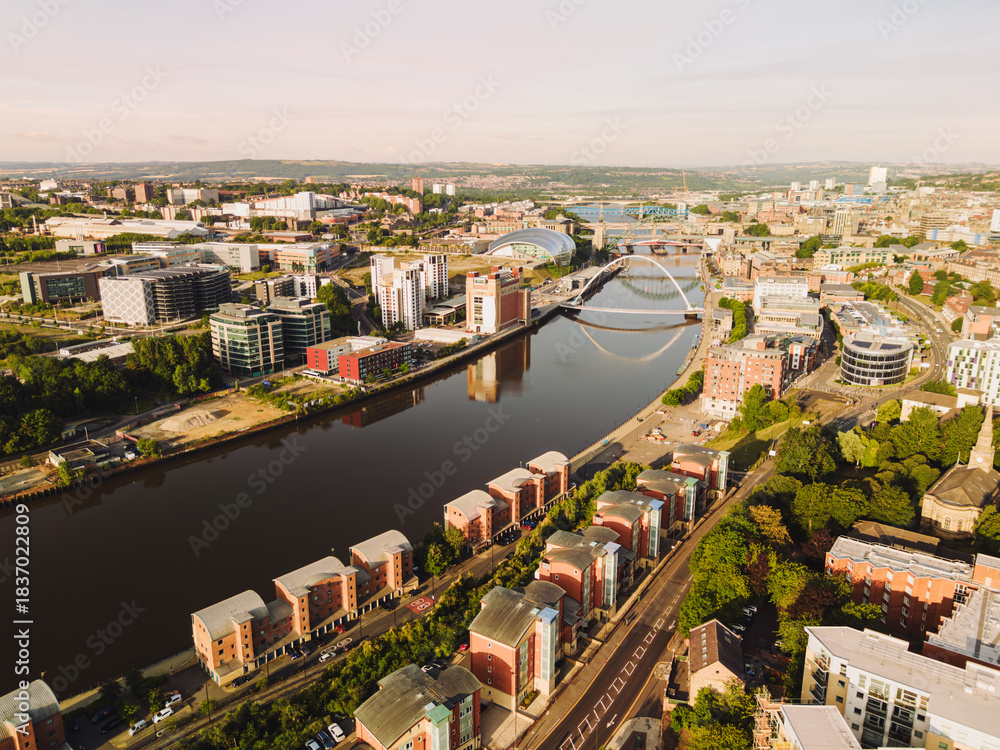 Obraz premium Newcastle upon Tyne: 9th August 2025: Buildings line the Newcastle Quayside at dawn. Bridges stands over the river, reflecting in the water below. Drone view from Ouseburn