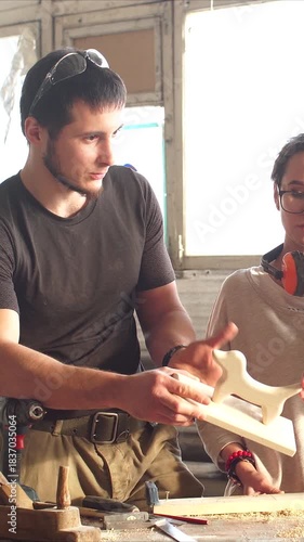 Man and woman in workshop inspecting carved wooden model. Instructor explains technique on carpentry lesson, sunlight through windows. Vertical video