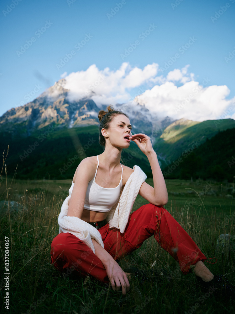 Obraz premium Outdoor landscape scene with a model seated in a grassy field, mountains in the distance, wearing a white crop top and red pants, a towel over shoulders, capturing a calm, nature infused moment.