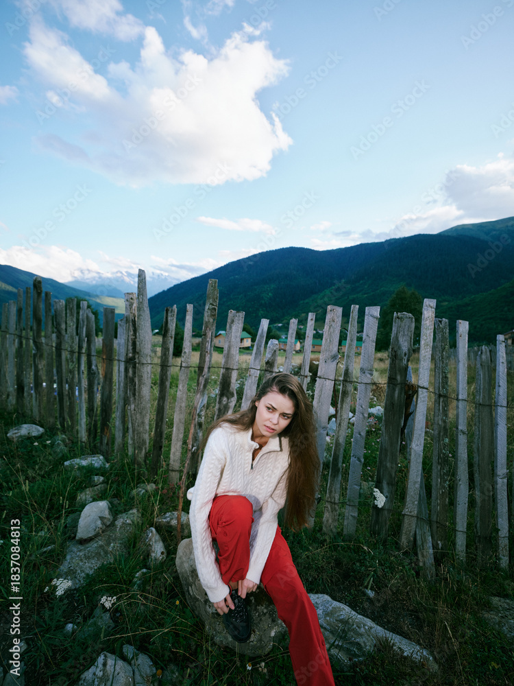 Obraz premium Woman sits by a weathered wooden fence in a mountainous landscape, wearing a light sweater and red pants, exploring nature and countryside ambience under a blue sky with scattered clouds.