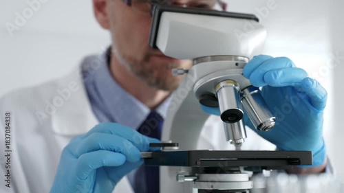 Close up of male scientist in white coat and blue gloves adjusting microscope, conducting pharmaceutical research in modern laboratory. Medicine and science concept