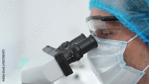 Masked scientist examining sample through microscope, wearing protective lab gear during medical research. Medicine, healthcare and science concept