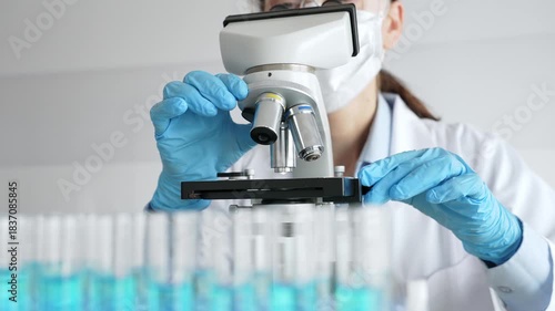 Female scientist wearing protective equipment checking microscope, analyzing blue test tubes in modern laboratory research setting. Medicine, healthcare and science concept