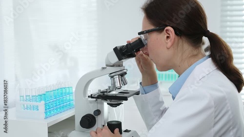 Female scientist wearing protective gear analyzing test samples under microscope in contemporary research environment. Medicine, healthcare and science concept