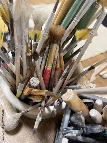 A dense collection of various art brushes (fan, round, flat) with wooden and metal handles, stored in a white container in a messy, creative art studio environment