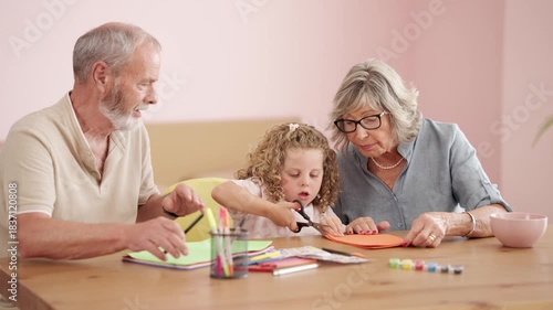 Loving grandparents spending quality time with their adorable granddaughter, teaching her how to draw and cut paper at a wooden table
