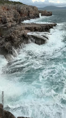 Powerful turquoise ocean waves crash against the rugged rocky shore. Turbulent seawater laps against the rocks during a storm