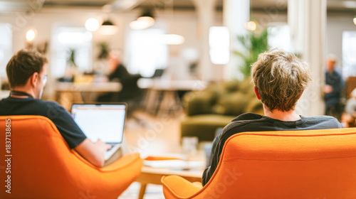 Two people sit in orange chairs in a modern office space, focused on their laptops, surrounded by a bright and inviting atmosphere.