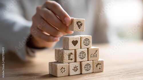 a person stacking wooden cubes into a pyramid shape, each cube has an icon: heart, gear, graph, globe, on a light wood table, natural daylight