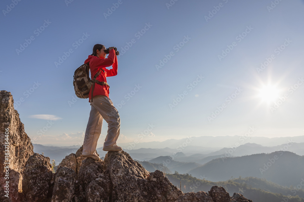 Naklejka premium Cute girl hiker with backpack holding binoculars standing on top of the rock mountain sunset sky background