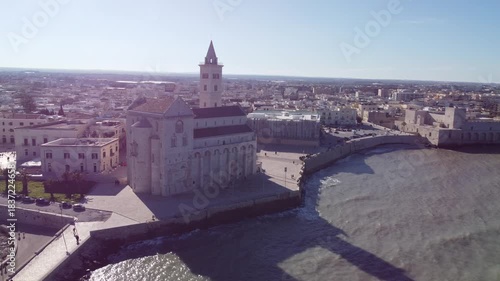 A panoramic coastal view of Trani shows the famous seaside cathedral overlooking turquoise waters, a long breakwater with lighthouses, and the historic old town bathed in bright Mediterranean sunlight