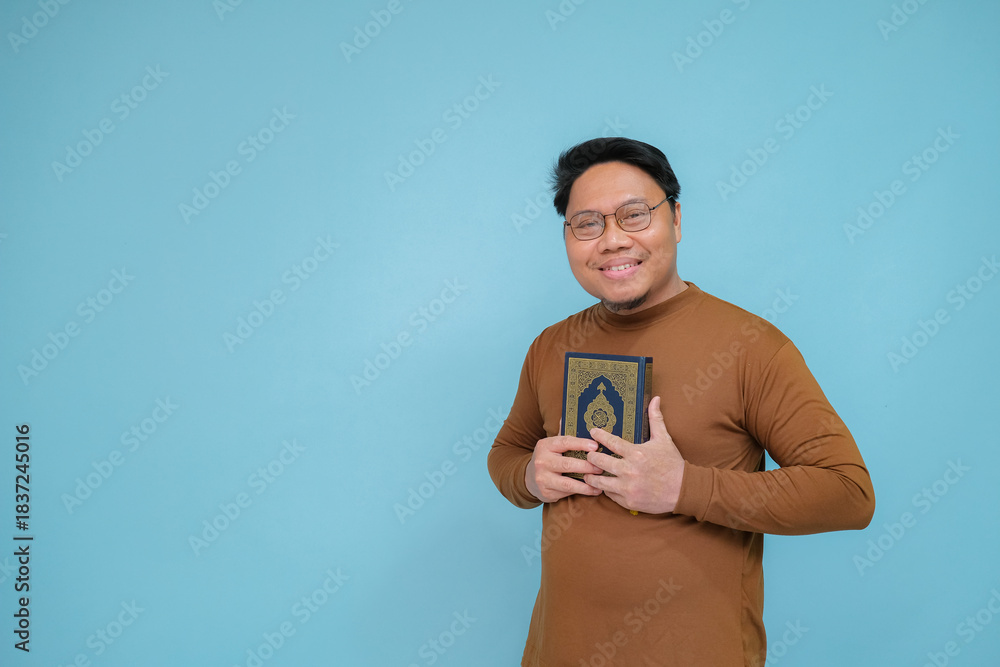 Fototapeta premium Portrait of an Asian Muslim man embracing the holy book of the Koran wearing brown shirt and glasses, the holy book of the Koran is guide for life.