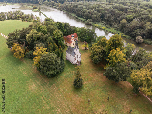 Fototapeta Naklejka Na Ścianę i Meble -  Historische Kapelle und Kirche am Flussufer, umgeben von Wald und Wiesen – Luftaufnahme
