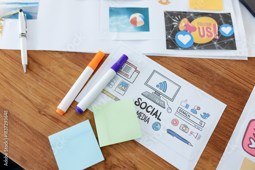 Sheet featuring SOCIAL MEDIA icons is sitting on wooden desk with markers and sticky notes nearby