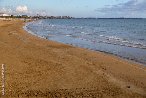 Fototapeta Naklejka Na Ścianę i Meble -  A wide, empty beach with wet sand and gentle surf under warm evening light, silhouettes of people and distant hotels visible on the calm coastline. Evrenseki, Antalya, Turkey, Mediterranean.
