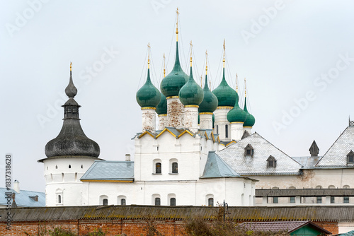 An autumn view of the green domes of the Rostov Kremlin from the outside. The white stone walls of the cathedral stand in front of a brick wall.