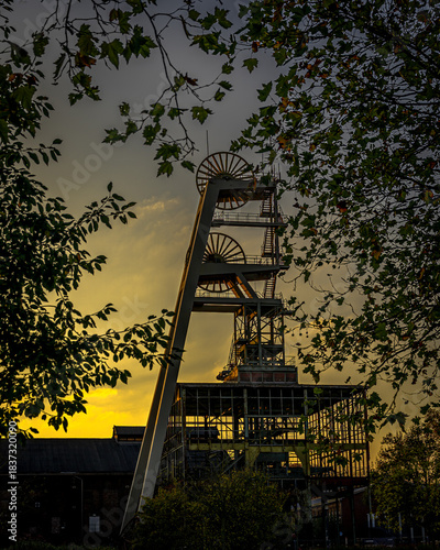 Das filigrane Stahlgerüst des Förderturms, eine Kathedrale der Ingenieurskunst, ragt als dunkle Silhouette in einen Himmel, der in tiefem Gelb und Orange glüht. Die mächtigen Seilräder sind die stumme
