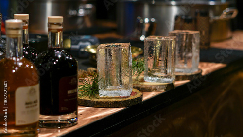 A rustic bar scene featuring whiskey bottles beside drinking glasses made from blocks of ice on a wooden counter. Christmas market decorations, festive shopping.