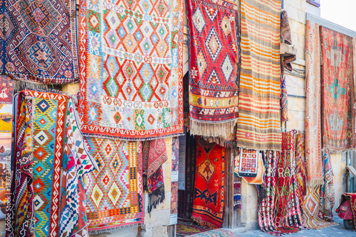 A vibrant display of traditional Turkish kilim rugs hanging outside a market shop in Cappadocia. The richly woven geometric patterns, bold colors, and diverse textile designs
