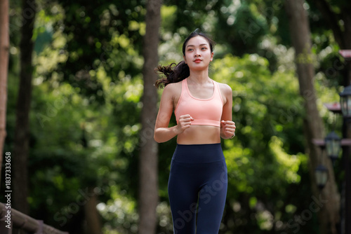 Young Asian woman wearing sports bra and leggings, running outdoors through a green park, promoting fitness and healthy lifestyle