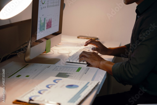 Man crunching numbers and analyzing charts on a computer in a dimly lit office, demonstrating dedication and overtime work
