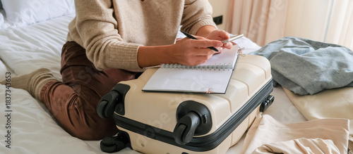 Travel planning scene with carry-on suitcase used as desk, open notebook and smartphone, hands writing a packing list in a cozy hotel room, minimalist weekend getaway prep, warm light lifestyle.