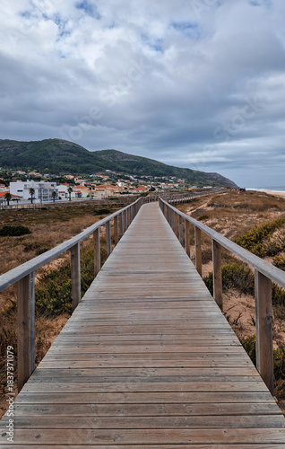 Long wooden boardwalk extending along a sandy dune landscape to a coastal town and hills under an overcast sky in Quiaios, Portugal