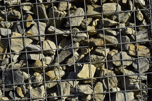 Gabion wall made of metal mesh filled with natural stones of different shapes and colors showing rough texture and contrast shadows