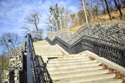 Concrete outdoor stairs with metal railings between gabion stone walls and leafless autumn trees under a bright blue sky