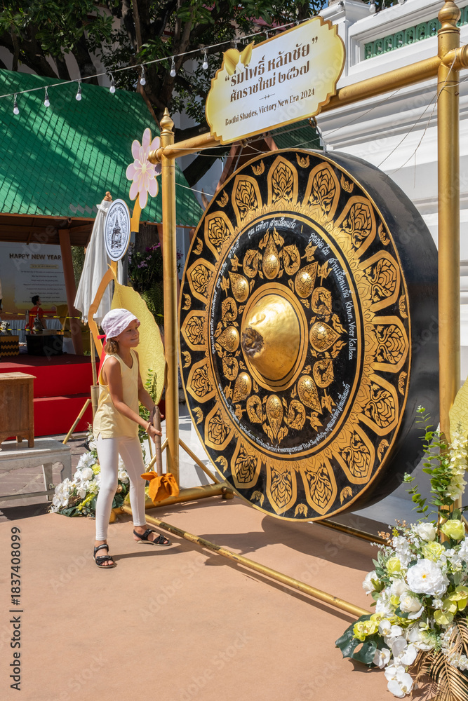 Naklejka premium A girl stands in front of a large gong at Wat Pho temple in Bangkok. She raises a mallet to strike the gong while celebrating New Year