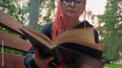 lady reading outdoors, girl with colorful dreadlocks focused on reading in quiet park setting, woman sporting pink dreadlocks quietly immersed in reading in tranquil outdoor environment