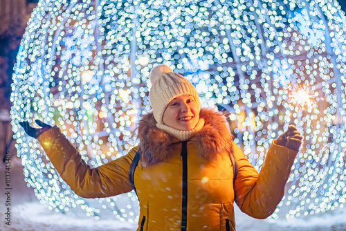 Young woman in knitted hat holds sparkler in her hands. Night festive street, shiny city decorations for Christmas.