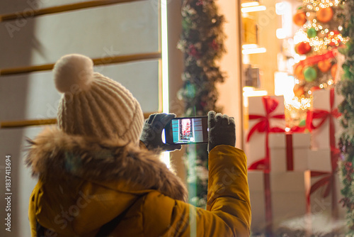 Woman tourist in knitted hat takes photographs of New Year's festive city decor on her smartphone. Christmas market, night lights. View from the back.