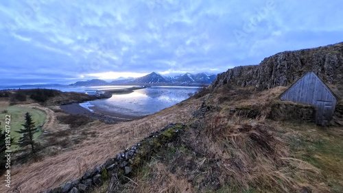 A wide, panoramic view over a grassy, rugged coastal hillside reveals a calm bay reflecting a cloudy, dramatic sky, with snow-capped mountains visible in the distant background near Borganes, Iceland