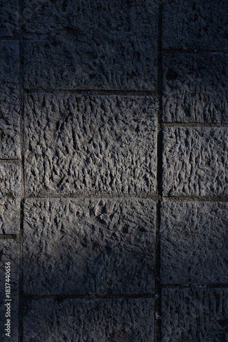A dark, moody photograph of chiseled stone tiles arranged in a grid-like structure. Subtle directional light highlights the carved texture and variations in each slab, emphasizing craftsmanship. 