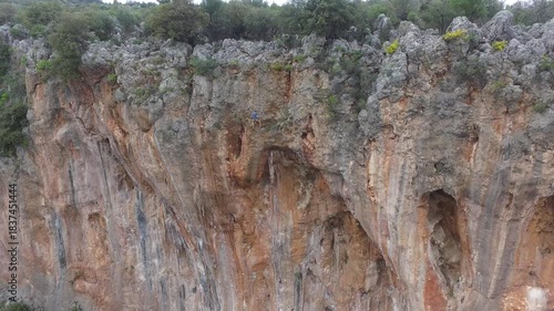 Aerial view of mountaineers rock climbing on a high natural rocky wall in rugged mountain terrain. Climbers secured by rope hang on a long karst cliff face as they ascend a steep wilderness formation.