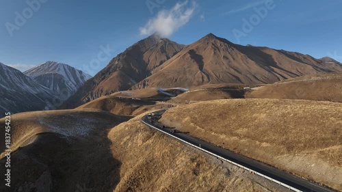 Aerial view of the winding Georgian Military Highway near Gudauri, Georgia. Mountain road surrounded by golden slopes and snowy peaks in early winter.