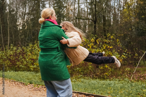 Spinning mother with daughter on park path. Playful hug shows joy, love, active childhood. Outdoor family game supports bonding, trust, balance, motion, energy, healthy lifestyle, weekend fun.