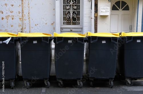 Fototapeta Naklejka Na Ścianę i Meble -  Row of black trash bins with yellow lids on street of small city. Waste management and rubbish sorting. Municipal management in european town