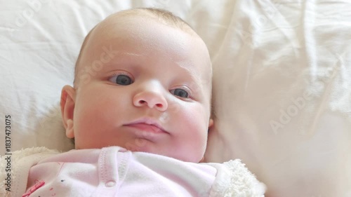 A baby is lying on a bed, wearing a pink shirt. The baby's face is visible, and it is smiling. The bed is covered in white sheets, and the room is well-lit