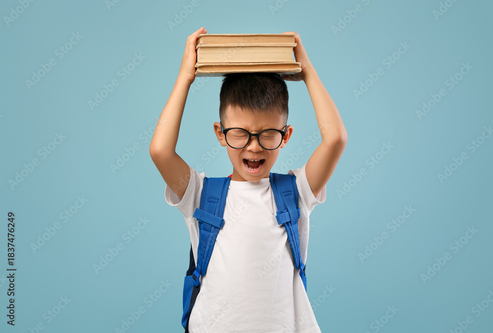 Fototapeta premium Young boy wearing glasses and a backpack holds a stack of books above his head with a frustrated expression, illustrating the challenge of carrying school materials.