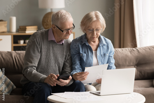 Domestic accounting in retirement. Busy older family couple engaged in finance paperwork at cozy home office check bills with calculator to pay online via notebook discuss expenses plan budget economy