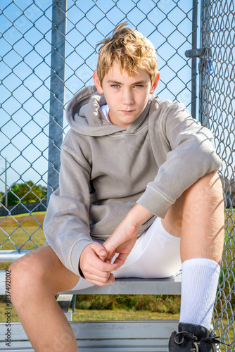 Young blonde boy in a grey hoodie sitting against a chain link fence on bleachers waiting for a game to start