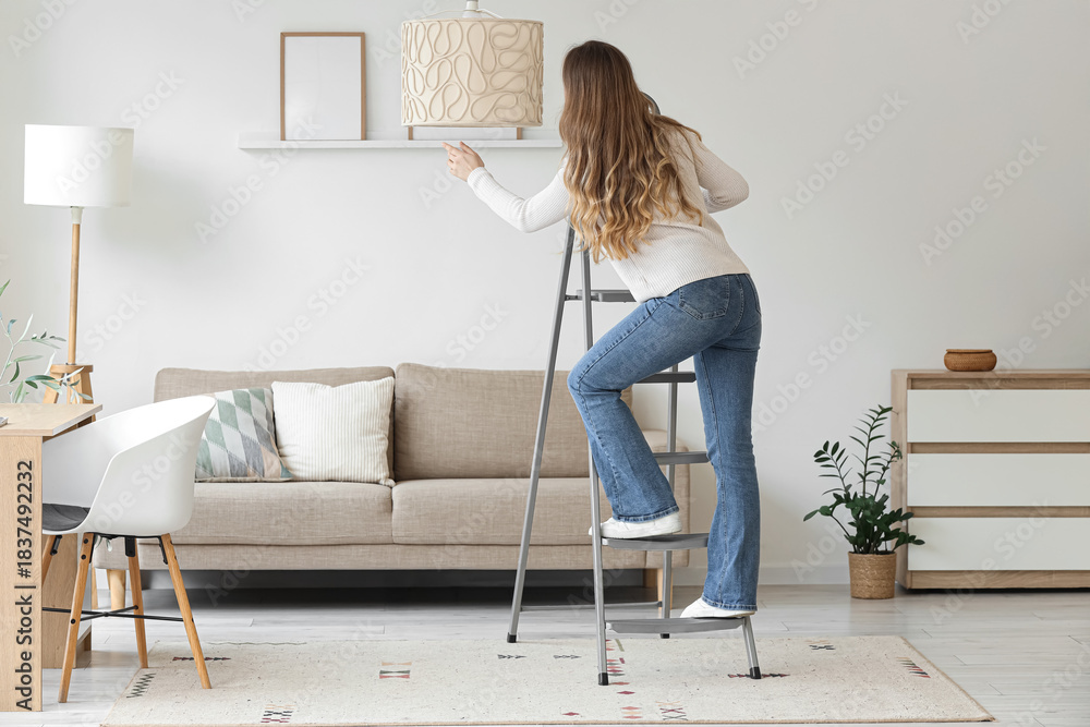 Obraz premium Young woman climbing stepladder near lamp in living room, back view